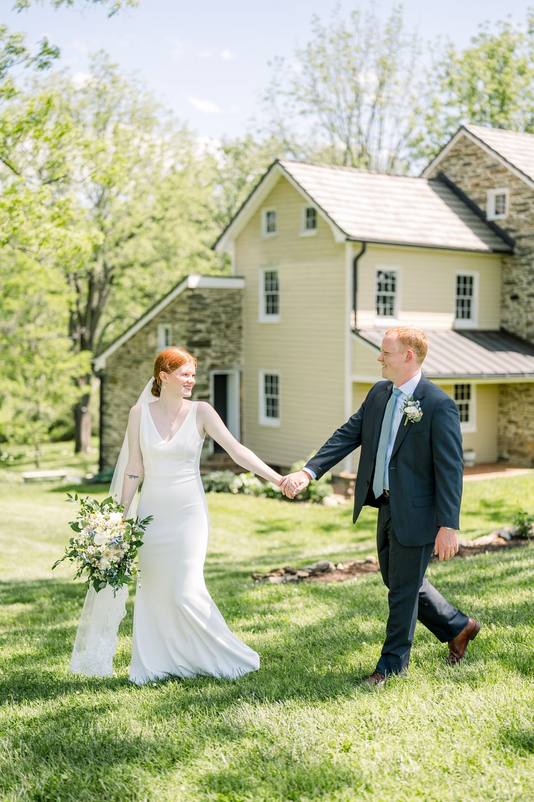 bride and groom holding hands and walking with stone farmhouse behind them for this Dreamy Spring Backyard Wedding